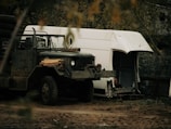 A rugged military-style truck with large tires is parked in a natural, rustic setting. It has a worn, weathered appearance and carries some lumber on its hood. Behind the truck, there is a partially dismantled white van or trailer with its side open, suggesting disrepair or modification. The background is filled with trees and foliage, creating an overgrown, secluded atmosphere.