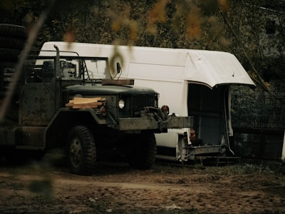 A rugged military-style truck with large tires is parked in a natural, rustic setting. It has a worn, weathered appearance and carries some lumber on its hood. Behind the truck, there is a partially dismantled white van or trailer with its side open, suggesting disrepair or modification. The background is filled with trees and foliage, creating an overgrown, secluded atmosphere.