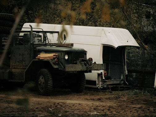 A rugged military-style truck with large tires is parked in a natural, rustic setting. It has a worn, weathered appearance and carries some lumber on its hood. Behind the truck, there is a partially dismantled white van or trailer with its side open, suggesting disrepair or modification. The background is filled with trees and foliage, creating an overgrown, secluded atmosphere.