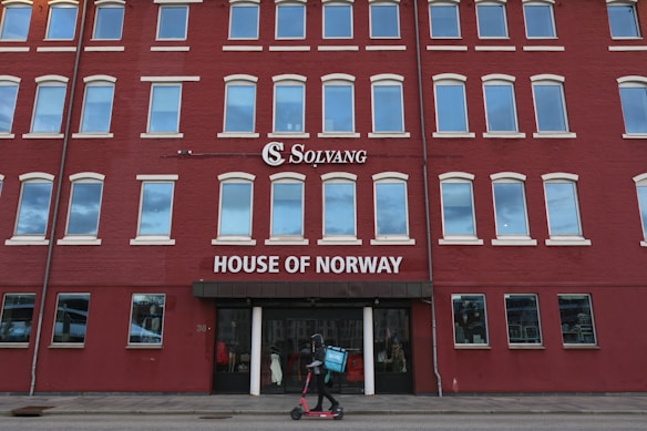 A multi-story red brick building with numerous white-framed windows. The facade displays the words 'Solvang' and 'House of Norway' in white lettering. A person rides an electric scooter along the sidewalk in front of the building, carrying a blue bag with a white logo.