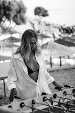 A woman with long hair is enjoying a game of foosball outdoors. She is wearing a light-colored shirt over a black bikini top. In the background, there are blurred images of trees and parasols, suggesting a beachside setting.