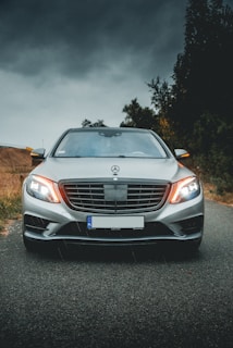 A silver luxury car is parked in the center of a road surrounded by greenery. The sky is overcast, creating a dramatic backdrop, and the car's headlights are on.