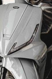 A close-up view of a sleek, modern scooter with a glossy, light gray finish. The design features sharp angles with integrated black accents resembling eyelids and a prominent brand logo. The front wheel and suspension are partially visible at the bottom.