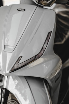 A close-up view of a sleek, modern scooter with a glossy, light gray finish. The design features sharp angles with integrated black accents resembling eyelids and a prominent brand logo. The front wheel and suspension are partially visible at the bottom.