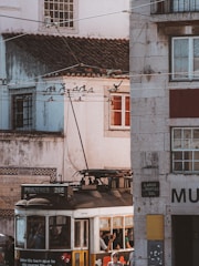 An old-fashioned tram passes through a narrow street lined with historic buildings. The tram is labeled 'PRAZERES 28E' and is bustling with passengers. Overhead tram wires crisscross above, and the building façades display signs of age with worn textures and a mix of colors. Sunlight casts warm tones on the scene, and a few people inside the tram are visible through the windows.