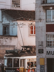 An old-fashioned tram passes through a narrow street lined with historic buildings. The tram is labeled 'PRAZERES 28E' and is bustling with passengers. Overhead tram wires crisscross above, and the building façades display signs of age with worn textures and a mix of colors. Sunlight casts warm tones on the scene, and a few people inside the tram are visible through the windows.