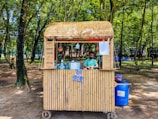 A small bamboo food stall with a straw roof is set in a lush forested area. A man stands behind the counter, preparing food or drink, with large metal pots and utensils visible. The surroundings are shaded by numerous trees and the ground is covered with dirt. Blue plastic chairs are scattered in the background.
