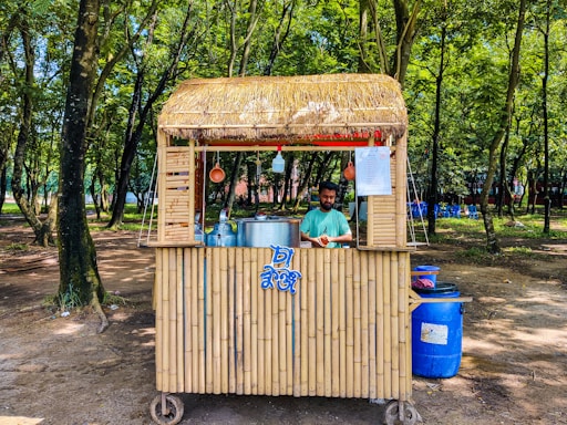 A small bamboo food stall with a straw roof is set in a lush forested area. A man stands behind the counter, preparing food or drink, with large metal pots and utensils visible. The surroundings are shaded by numerous trees and the ground is covered with dirt. Blue plastic chairs are scattered in the background.