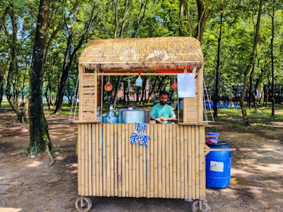 A small bamboo food stall with a straw roof is set in a lush forested area. A man stands behind the counter, preparing food or drink, with large metal pots and utensils visible. The surroundings are shaded by numerous trees and the ground is covered with dirt. Blue plastic chairs are scattered in the background.