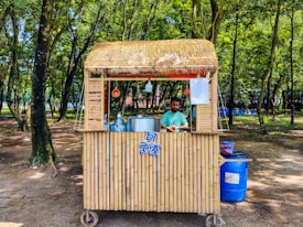 A small bamboo food stall with a straw roof is set in a lush forested area. A man stands behind the counter, preparing food or drink, with large metal pots and utensils visible. The surroundings are shaded by numerous trees and the ground is covered with dirt. Blue plastic chairs are scattered in the background.
