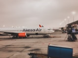 An airplane parked at an airport terminal with the Akasa Air logo visible on the fuselage. The aircraft is positioned on a tarmac with various equipment and vehicles nearby. Another airplane with a SpiceJet logo can be seen in the background. The sky appears overcast, indicating a cloudy weather condition.
