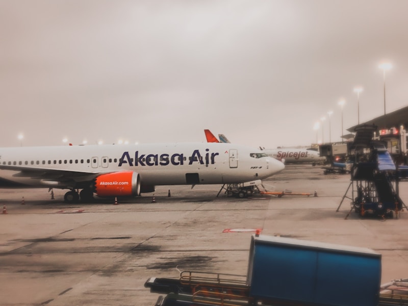 An airplane parked at an airport terminal with the Akasa Air logo visible on the fuselage. The aircraft is positioned on a tarmac with various equipment and vehicles nearby. Another airplane with a SpiceJet logo can be seen in the background. The sky appears overcast, indicating a cloudy weather condition.