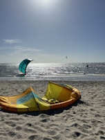 A sunny beach scene with kitesurfing equipment lying on the sand, including a deflated yellow and orange kite. In the background, people are kitesurfing on the water against the bright sky.