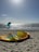 A sunny beach scene with kitesurfing equipment lying on the sand, including a deflated yellow and orange kite. In the background, people are kitesurfing on the water against the bright sky.