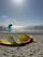 A sunny beach scene with kitesurfing equipment lying on the sand, including a deflated yellow and orange kite. In the background, people are kitesurfing on the water against the bright sky.
