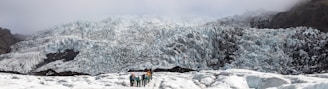 A group of people is hiking on a glacier with a massive ice formation towering in the background. The glacier is covered in rugged, jagged ice that appears bluish-white, and the hikers are dressed in colorful outdoor gear.