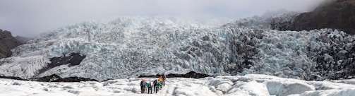 A group of people is hiking on a glacier with a massive ice formation towering in the background. The glacier is covered in rugged, jagged ice that appears bluish-white, and the hikers are dressed in colorful outdoor gear.