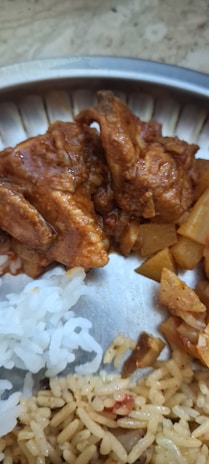A close-up of a stainless steel plate containing a variety of foods. There are pieces of curry chicken, cubes of potato curry, white rice, and a serving of seasoned rice, all arranged side by side on the plate.