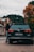 A person stands next to a black Volkswagen Passat with visible license plate on a paved street. The background includes autumnal trees with orange and green foliage, and a few old buildings along a tram line. The overcast sky suggests a cloudy day.