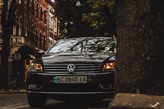 A dark-colored Volkswagen car is parked on a cobblestone street, partially obscured by a large tree. The vehicle's headlights are on, and the license plate is visible. Surrounding buildings are styled in an older European architectural design, with detailed facades and a warm ambiance created by the streetlights and shadows of nearby trees.