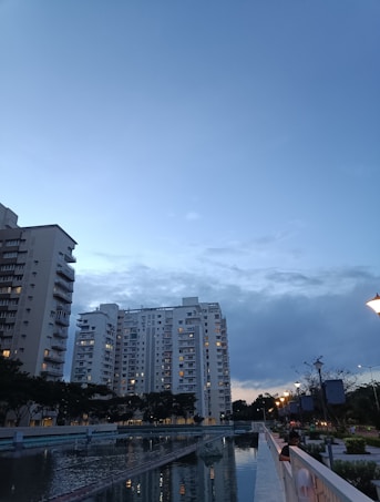 High-rise apartment buildings stand against a backdrop of a darkening evening sky. Some windows are lit, suggesting occupancy. A calm, reflective water body runs parallel to a walkway lined with streetlamps, casting a warm glow. Trees are visible along the walkway, contributing to the tranquil ambiance.