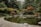 A serene Japanese garden featuring a calm pond surrounded by colorful autumn trees and lush greenery. A small waterfall flows into the pond, and the area is bordered by smooth stones. A traditional stone lantern is visible on the right side.