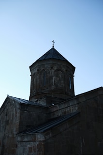 A stone church with a cylindrical tower topped by a small cross, set against a clear blue sky. The architectural style is simple yet historic, with weathered stones and a steep, dark roof.