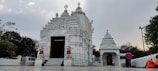 A beautiful white temple with intricate carvings stands against a cloudy sky. Two lion statues flank the entrance, which leads into a sanctum. People in colorful attire, including a figure in orange robes, are present around the temple.