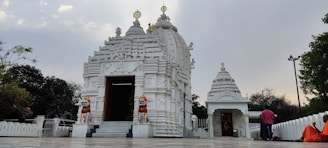 A beautiful white temple with intricate carvings stands against a cloudy sky. Two lion statues flank the entrance, which leads into a sanctum. People in colorful attire, including a figure in orange robes, are present around the temple.