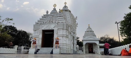 A beautiful white temple with intricate carvings stands against a cloudy sky. Two lion statues flank the entrance, which leads into a sanctum. People in colorful attire, including a figure in orange robes, are present around the temple.