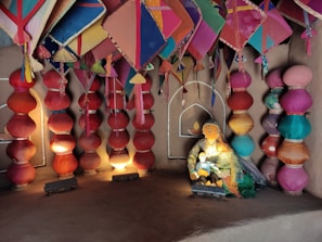 A rustic room decorated with colorful kites hanging from the ceiling and large, vibrant fabric lamps arranged in rows along the walls. A mannequin resembling a woman in traditional attire is sitting, while holding a child mannequin. A small painted alcove, resembling a candle or diya, adds to the room's cultural theme.