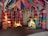A rustic room decorated with colorful kites hanging from the ceiling and large, vibrant fabric lamps arranged in rows along the walls. A mannequin resembling a woman in traditional attire is sitting, while holding a child mannequin. A small painted alcove, resembling a candle or diya, adds to the room's cultural theme.