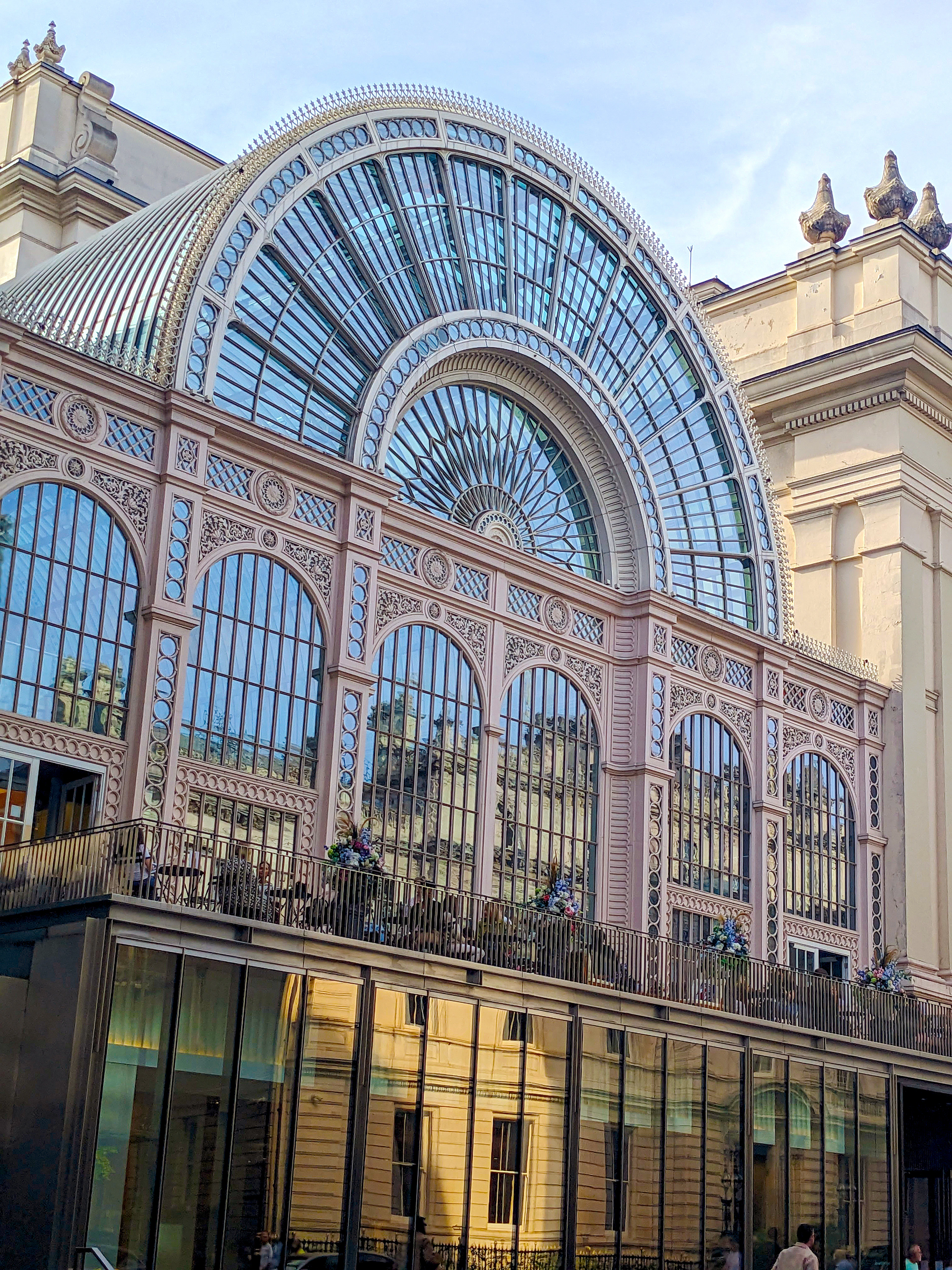 The glass and cast-iron facade of the Royal Opera House's Paul Hamlyn Hall in Covent Garden, its pale pink ironwork fanning into a great arched window above a balcony of flowering planters