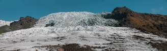 An expansive glacier sits against a backdrop of rocky, mountainous terrain. The glacier, composed of an intricate mass of ice, flows down from the mountains. There are patches of dark rock visible through the icy surface, with the surrounding mountains showing a stark contrast of rugged, brown rock against the white and blue ice. The sky above is clear with a rich blue color.