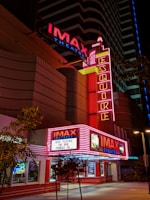 A nighttime view of an IMAX theater with bright red and pink neon lights illuminating the entrance. The marquee displays the titles 'The Nun' and 'Pandas 3D' as currently playing. The architecture is modern with vertical neon signs and additional advertisements visible. Trees are present near the entrance, contrasting with the urban setting.