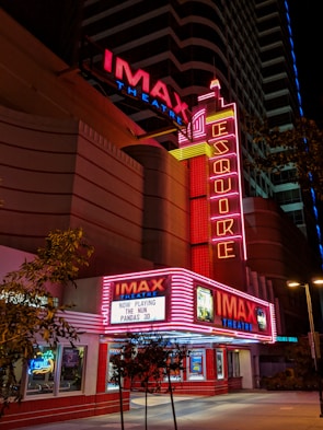 A nighttime view of an IMAX theater with bright red and pink neon lights illuminating the entrance. The marquee displays the titles 'The Nun' and 'Pandas 3D' as currently playing. The architecture is modern with vertical neon signs and additional advertisements visible. Trees are present near the entrance, contrasting with the urban setting.