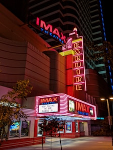 A nighttime view of an IMAX theater with bright red and pink neon lights illuminating the entrance. The marquee displays the titles 'The Nun' and 'Pandas 3D' as currently playing. The architecture is modern with vertical neon signs and additional advertisements visible. Trees are present near the entrance, contrasting with the urban setting.