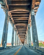 A perspective view of an empty roadway under a large concrete bridge, with visible support columns and beams creating distinct patterns. The sky is clear and blue, and some urban buildings can be seen in the background.