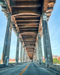 A perspective view of an empty roadway under a large concrete bridge, with visible support columns and beams creating distinct patterns. The sky is clear and blue, and some urban buildings can be seen in the background.