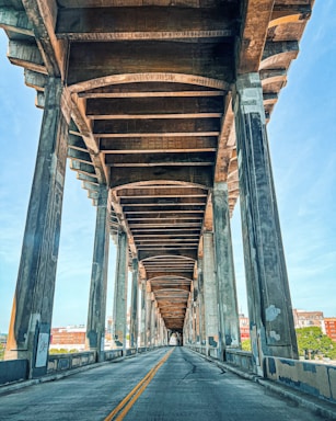 A perspective view of an empty roadway under a large concrete bridge, with visible support columns and beams creating distinct patterns. The sky is clear and blue, and some urban buildings can be seen in the background.