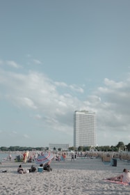 A sandy beach is crowded with people enjoying a sunny day. Families and groups are gathered under umbrellas. A tall hotel building is visible in the background, with a clear sky and a few fluffy clouds overhead.
