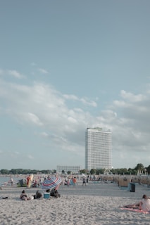 A sandy beach is crowded with people enjoying a sunny day. Families and groups are gathered under umbrellas. A tall hotel building is visible in the background, with a clear sky and a few fluffy clouds overhead.