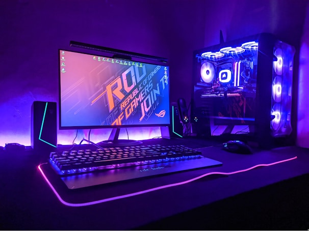 A gaming setup with a monitor displaying a colorful 'Republic of Gamers' logo, accompanied by a mechanical keyboard and a gaming mouse on a desk illuminated with RGB lighting. The computer tower is visible, featuring internal cooling fans lit with vibrant blue and purple LED lights.