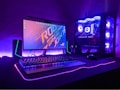 A gaming setup with a monitor displaying a colorful 'Republic of Gamers' logo, accompanied by a mechanical keyboard and a gaming mouse on a desk illuminated with RGB lighting. The computer tower is visible, featuring internal cooling fans lit with vibrant blue and purple LED lights.