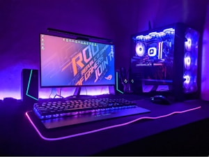 A gaming setup with a monitor displaying a colorful 'Republic of Gamers' logo, accompanied by a mechanical keyboard and a gaming mouse on a desk illuminated with RGB lighting. The computer tower is visible, featuring internal cooling fans lit with vibrant blue and purple LED lights.