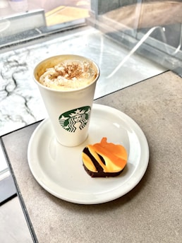 A cup of coffee topped with whipped cream and cinnamon is placed on a white saucer. Next to it is a small chocolate covered treat decorated with orange and black icing shaped like a pumpkin. The setting is inside a café, with a window showing a blurry outdoor scene.