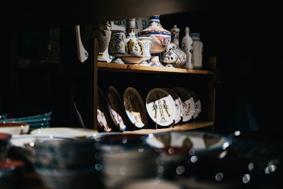 An assortment of decorative ceramic dishes and vases is displayed on wooden shelves in a dimly lit space. The ceramics feature intricate patterns and vibrant colors, with some showcasing floral designs. Light highlights the colorful patterns on the ceramics, drawing attention to their artistic details.