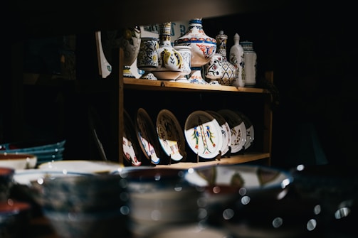 An assortment of decorative ceramic dishes and vases is displayed on wooden shelves in a dimly lit space. The ceramics feature intricate patterns and vibrant colors, with some showcasing floral designs. Light highlights the colorful patterns on the ceramics, drawing attention to their artistic details.