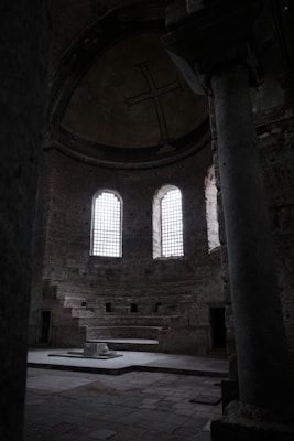 A dimly lit, ancient stone interior features high, arched windows with grid-like patterns that allow sunlight to filter through. The walls are made of weathered stone, and a large cross is prominently displayed on the upper wall. A series of ascending stone benches lies beneath, suggesting a former place of gathering or worship. The space is expansive and echoes with historical significance.