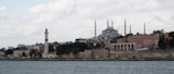 The image displays a coastal cityscape featuring a prominent mosque with multiple minarets, set against a cloudy sky. In the foreground, there is a shoreline with ancient stone walls, a lighthouse, and several historic buildings.